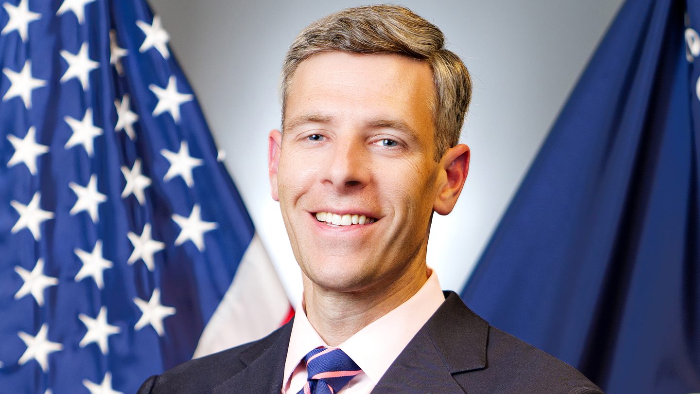 Official portrait of Comptroller of the Currency Jonathan Gould in front of US and OCC flags