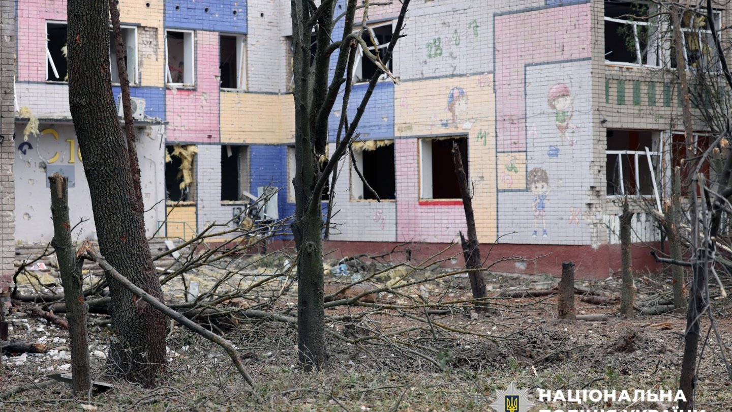 A damaged, colourfully painted multi-storey building with broken windows behind bare trees and debris in Kherson, Ukraine