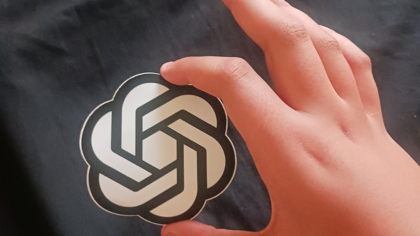 A hand holding a white OpenAI logo sticker against a dark fabric background