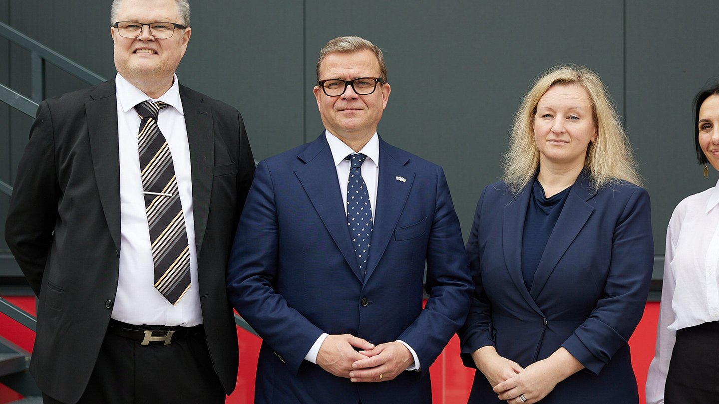 Finnish Prime Minister Petteri Orpo, in a dark suit and blue tie, stands with officials in front of a red wall