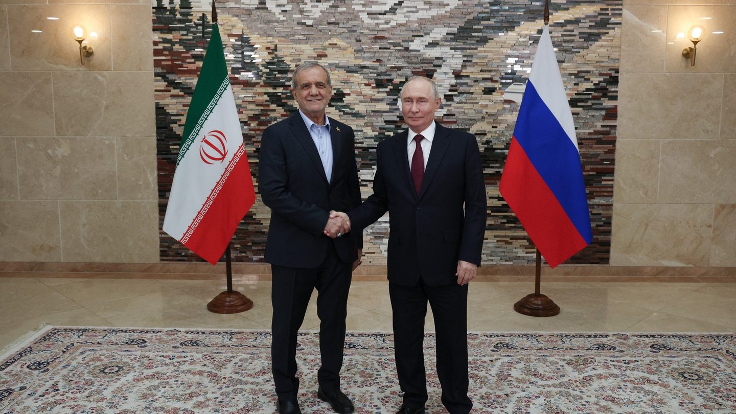 Iranian President Masoud Pezeshkian and Russian President Vladimir Putin shake hands in front of their national flags