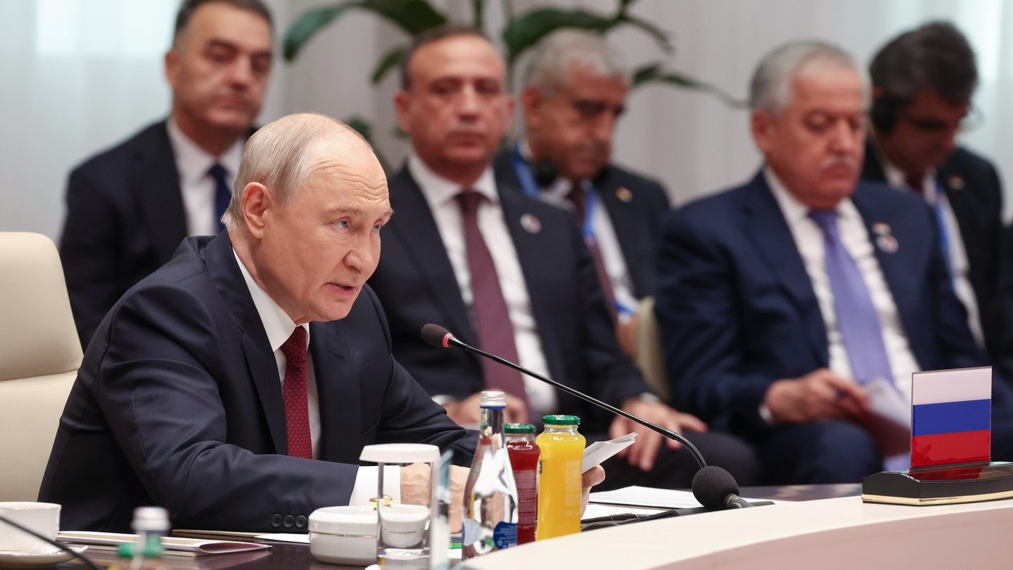 Russian President Vladimir Putin speaks into a microphone at a conference table, with delegates seated behind him and a Russian flag on the table
