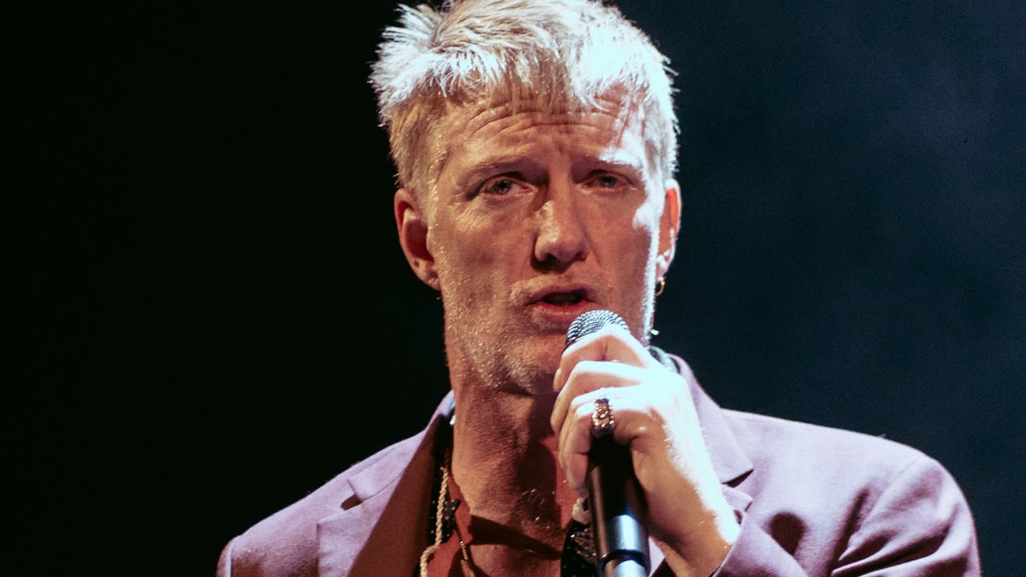 Queens of the Stone Age singer Josh Homme, with short blond hair and a maroon jacket, sings into a microphone on a dark stage