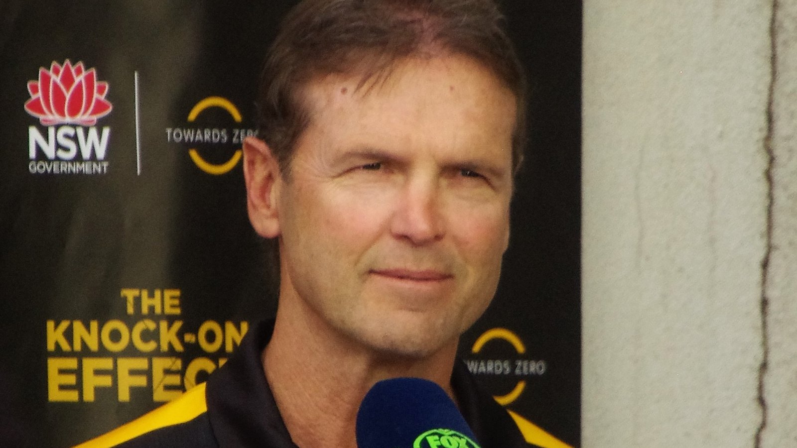 Mick Potter, wearing a black and yellow polo shirt, speaks into a microphone in front of a sponsor backdrop