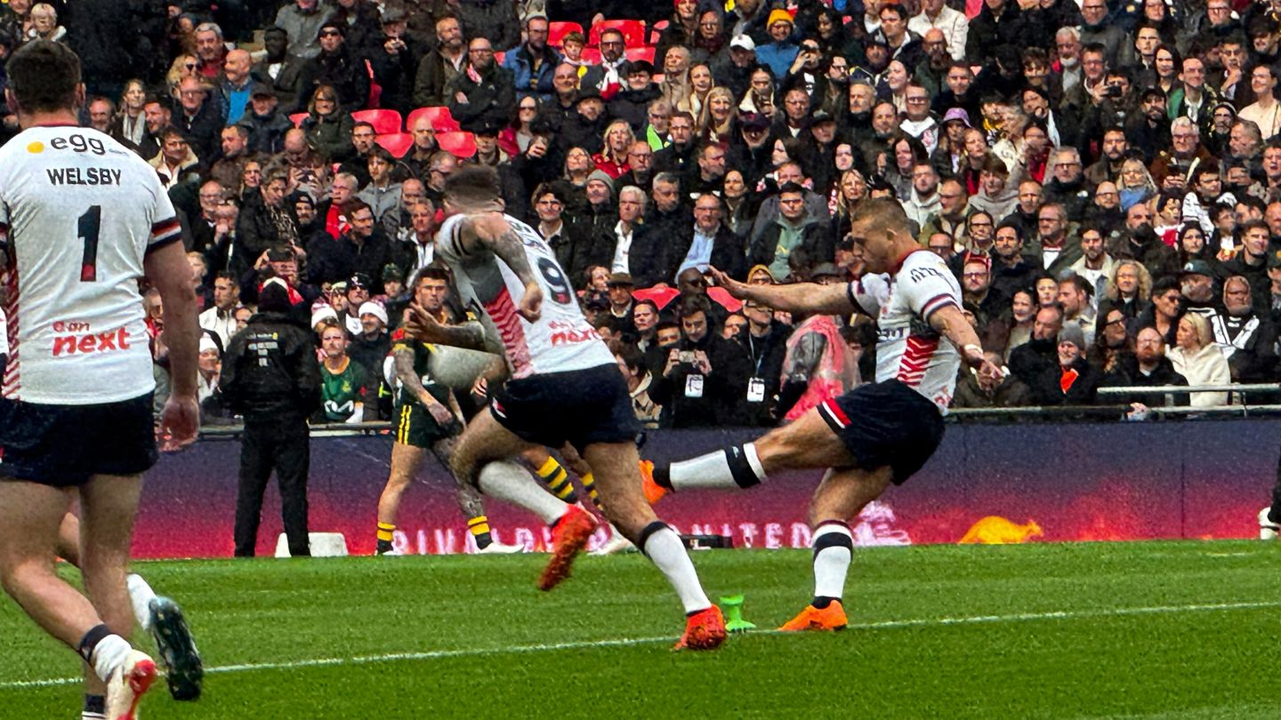 England rugby league player in a white kit kicks off during a Test match, with teammates including Jack Welsby running forward and a full crowd behind