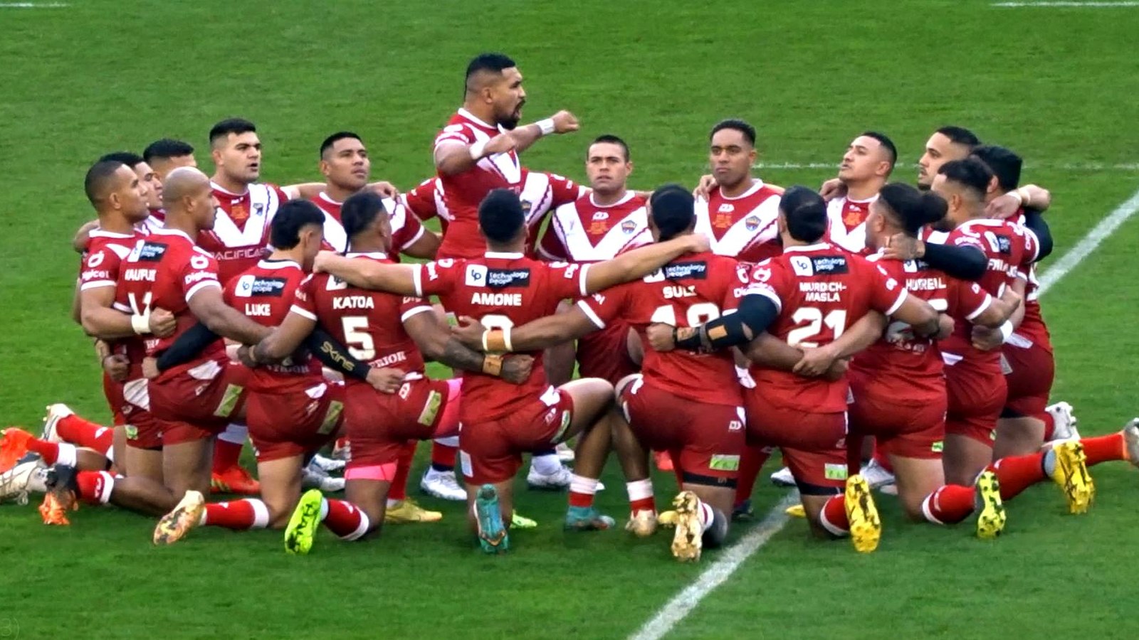 Tonga rugby league players in red and white uniforms gathered in a circle on the pitch performing the Sipi Tau before a World Cup match