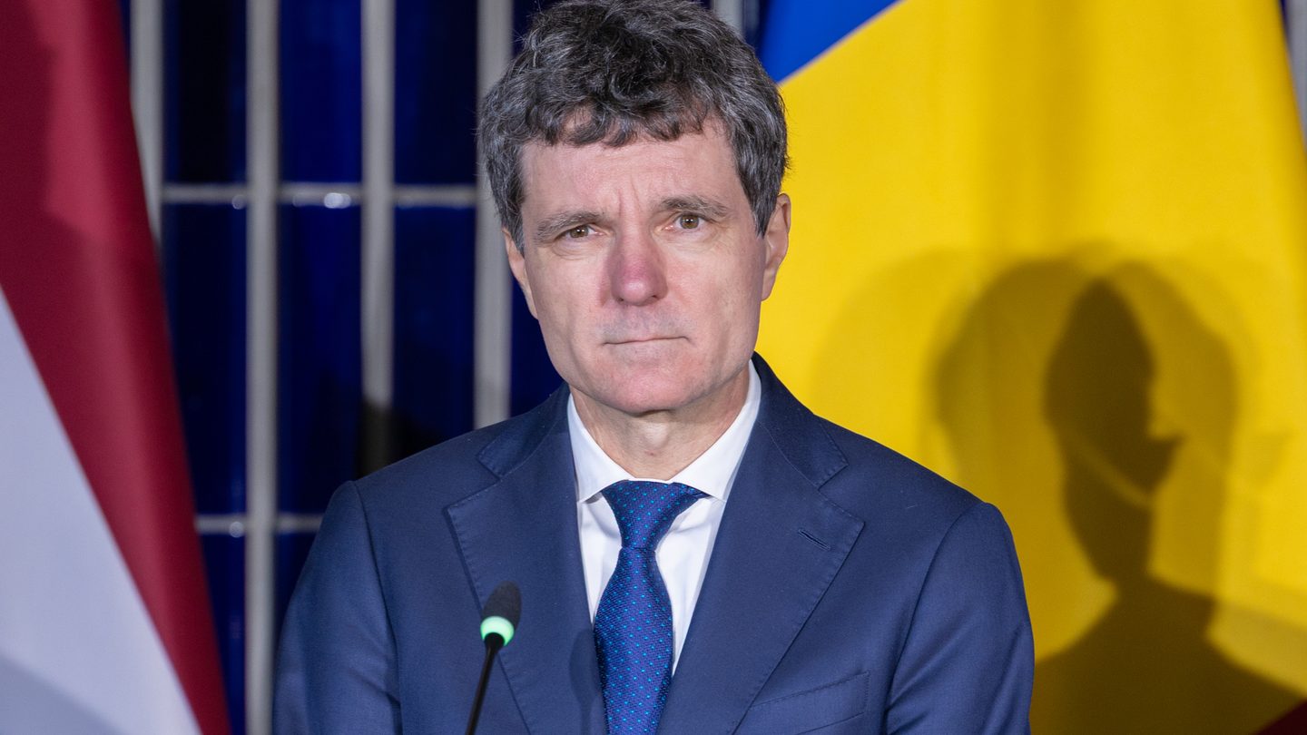 Romanian President Nicușor Dan, in a dark suit and blue tie, standing at a podium in front of flags