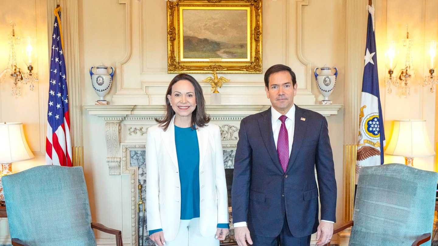 María Corina Machado, in a white jacket, stands beside US Secretary of State Marco Rubio in a formal room at the State Department with US flags