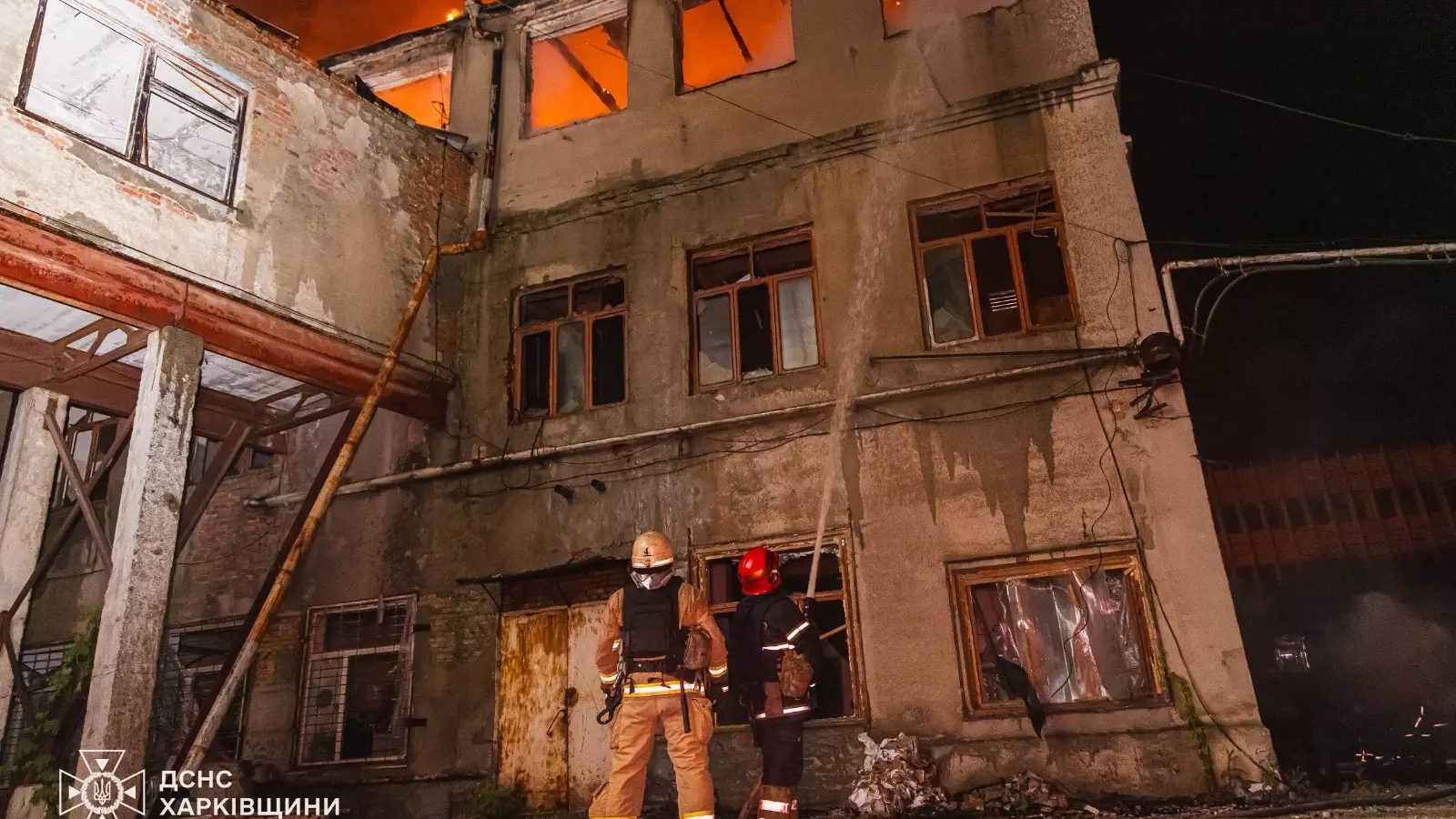 Two Ukrainian firefighters in helmets spray water at a damaged multi-storey industrial building with fire burning inside its upper windows at night in Kharkiv