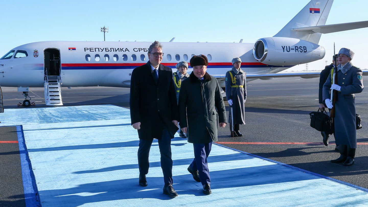 Aleksandar Vučić, in a dark coat and tie, walks along a blue carpet past an honour guard after leaving a white Serbian government jet