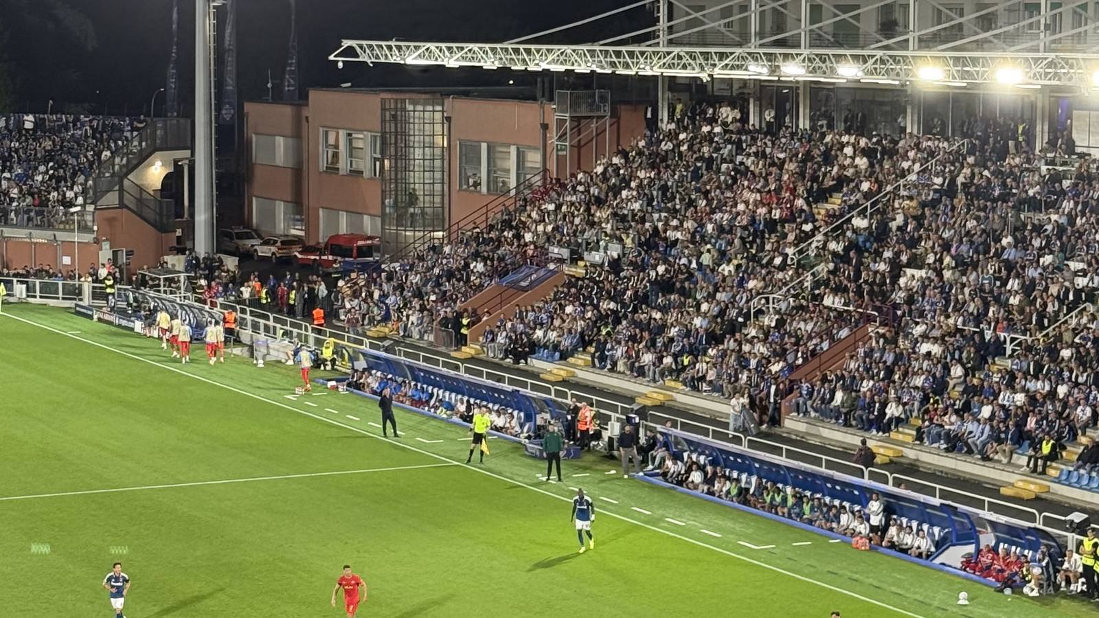 Night match at Como's Stadio Sinigaglia seen from behind the goal, with players on the floodlit pitch and a full stand opposite