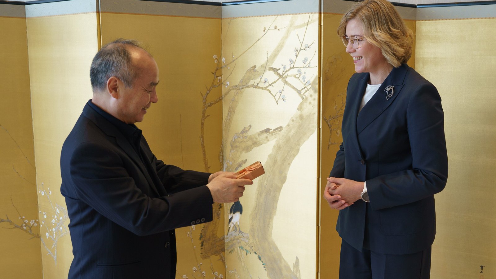 Masayoshi Son, left, exchanges a gift with European Commission Executive Vice-President Henna Virkkunen in front of a gold folding screen