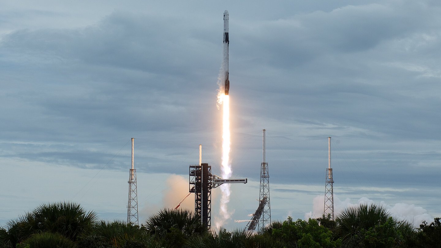 A SpaceX Falcon 9 rocket lifts off on a bright flame from a launch pad flanked by tall lightning towers, under a grey cloudy sky with palm trees in the foreground
