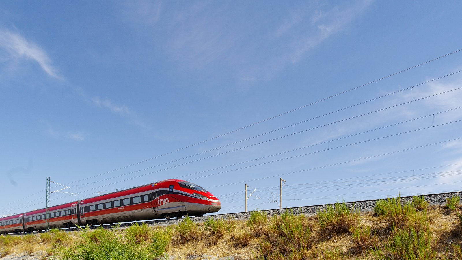 A red Iryo Frecciarossa 1000 high-speed train running on an electrified line near Madrid under a blue sky