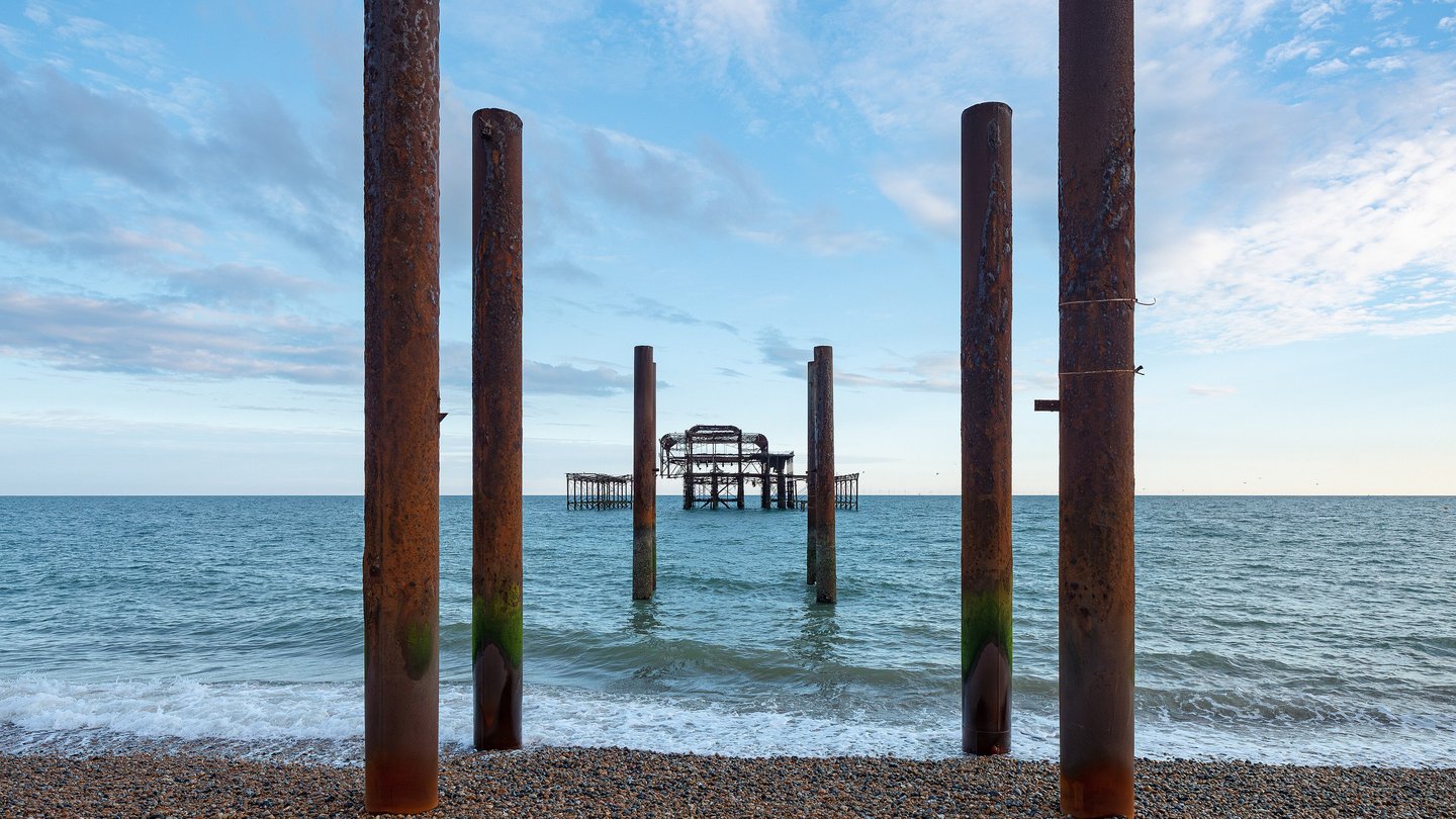 Rusted iron columns standing on a pebble beach in front of the skeletal remains of Brighton's West Pier out at sea under a blue sky