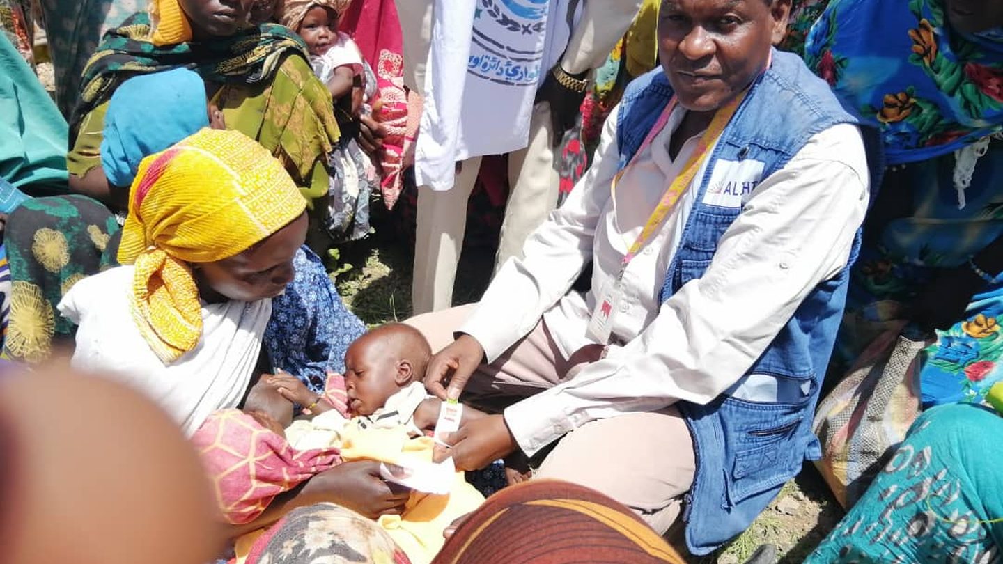 An aid worker in a blue vest checks a baby held by a woman during malnutrition screening in a village in Sudan, with other women waiting nearby