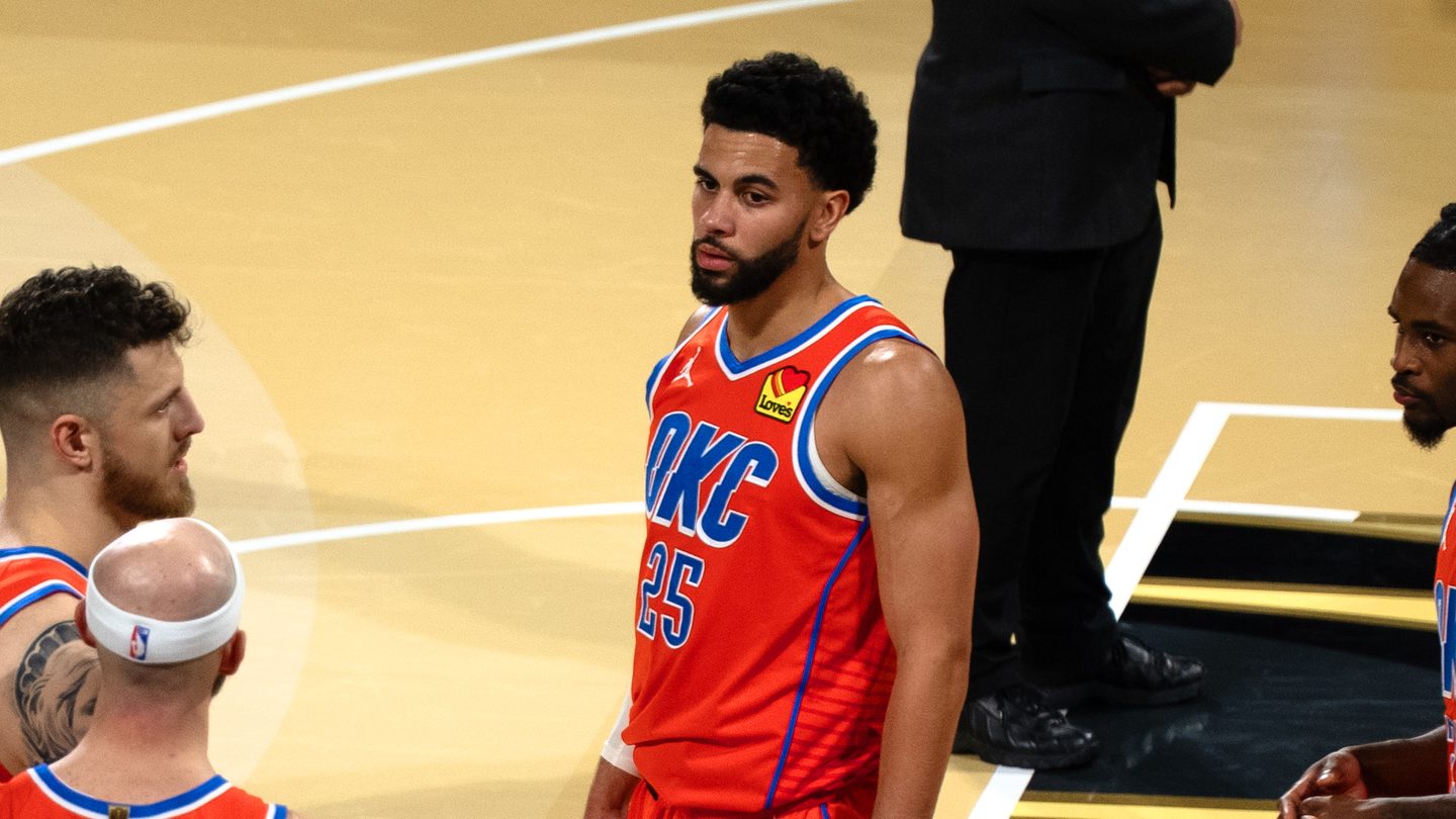 Oklahoma City Thunder guard Ajay Mitchell, in an orange number 25 OKC jersey, stands on court next to teammates Isaiah Hartenstein and Alex Caruso