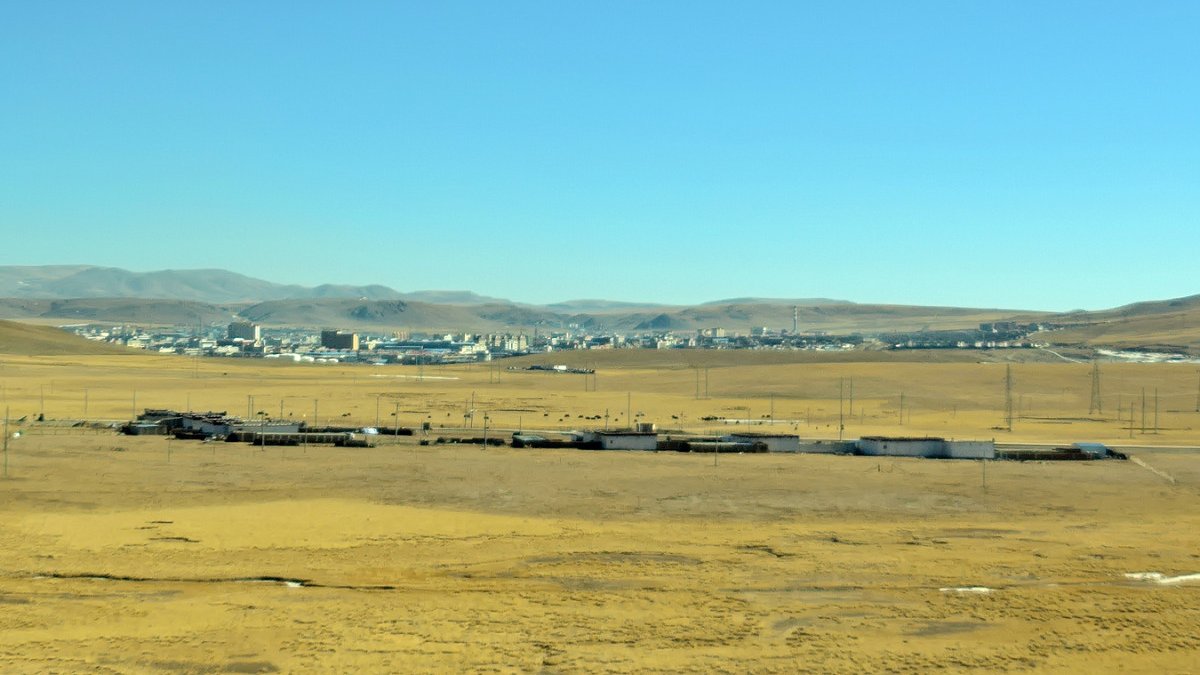 A wide grassland plateau with low hills and the buildings of Nagqu town in the distance under a clear blue sky