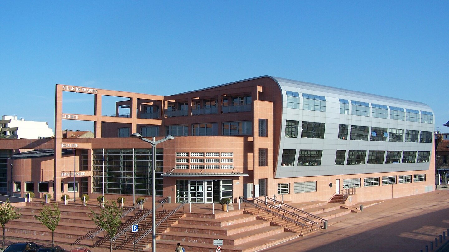 The modern brick and grey-metal town hall of Trappes, with 'Ville de Trappes' and the words Liberté, Égalité, Fraternité on its facade, under a blue sky