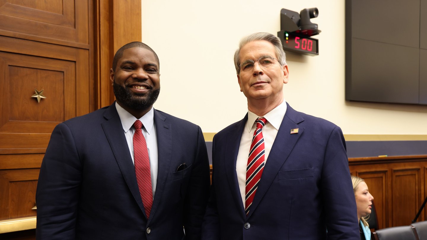 Treasury Secretary Scott Bessent, in a dark suit and striped red tie, stands beside Congressman Byron Donalds in a wood-panelled room