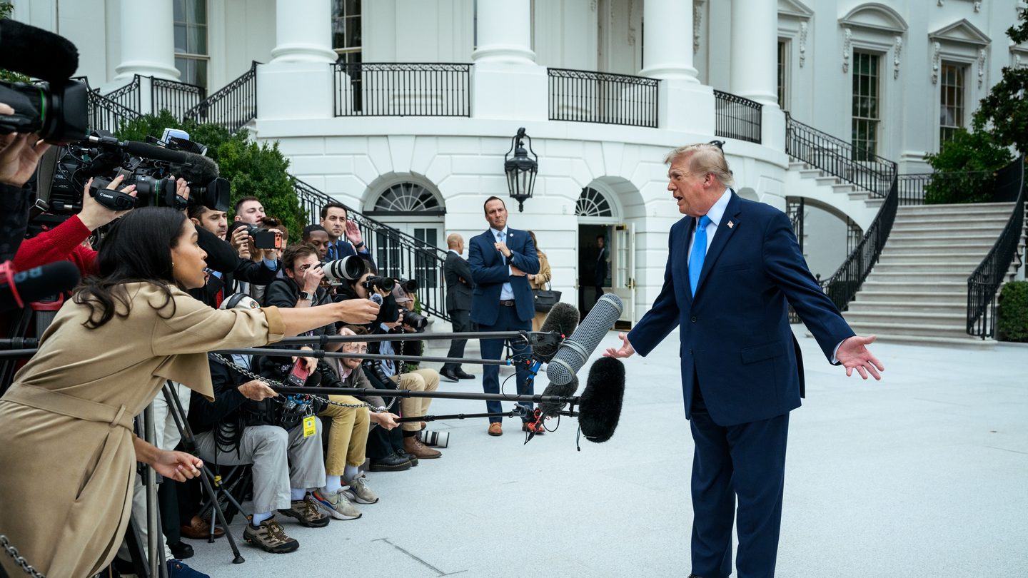 President Donald Trump, in a dark blue suit and light blue tie, gestures as he speaks to reporters holding microphones and cameras on the South Lawn of the White House