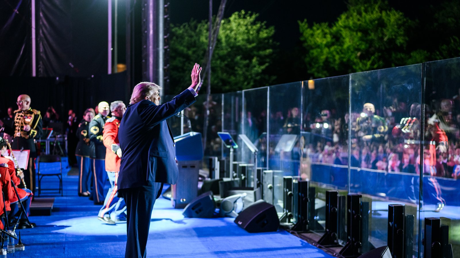 Donald Trump, in a dark suit, waves to a crowd from a blue-lit stage at an evening rally, with Marine Band musicians beside him