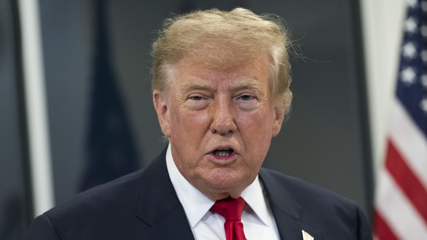 President Donald Trump, in a dark suit and red tie, speaks to reporters with a US flag behind him