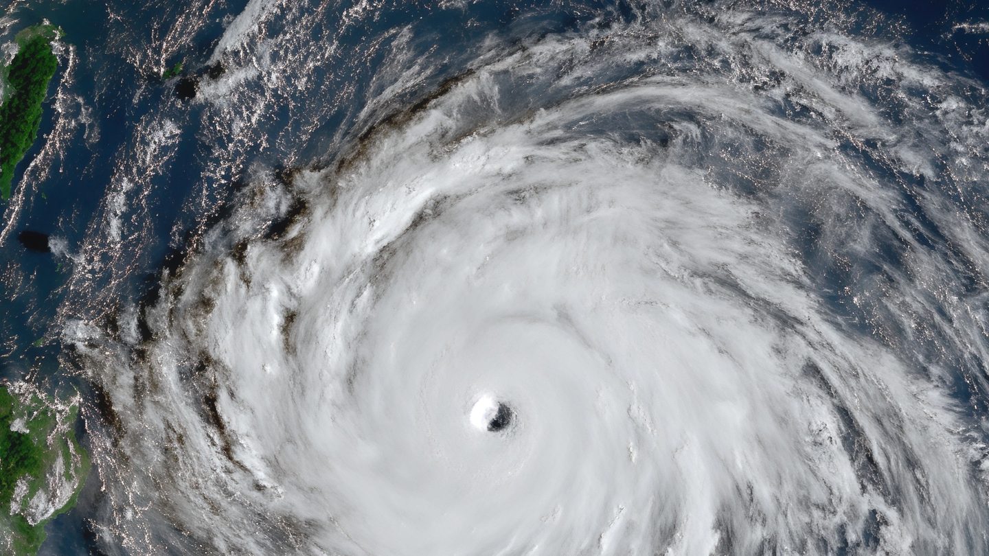Satellite view of a large spiral of white cloud with a small clear eye over a dark blue ocean, with green islands at the left edge
