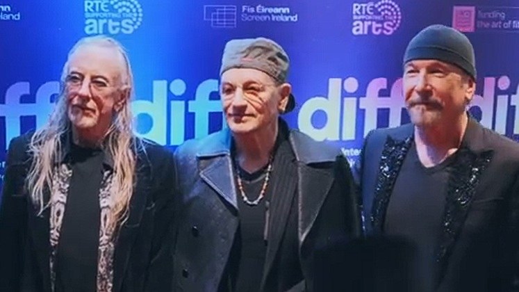Bono, in a grey cap and tinted glasses, and the Edge, in a black beanie, standing with the artist Guggi in front of a blue Dublin International Film Festival backdrop