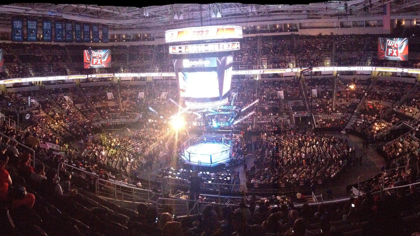 High-angle wide view of a packed arena with a lit UFC octagon in the centre, surrounded by crowds and big screens