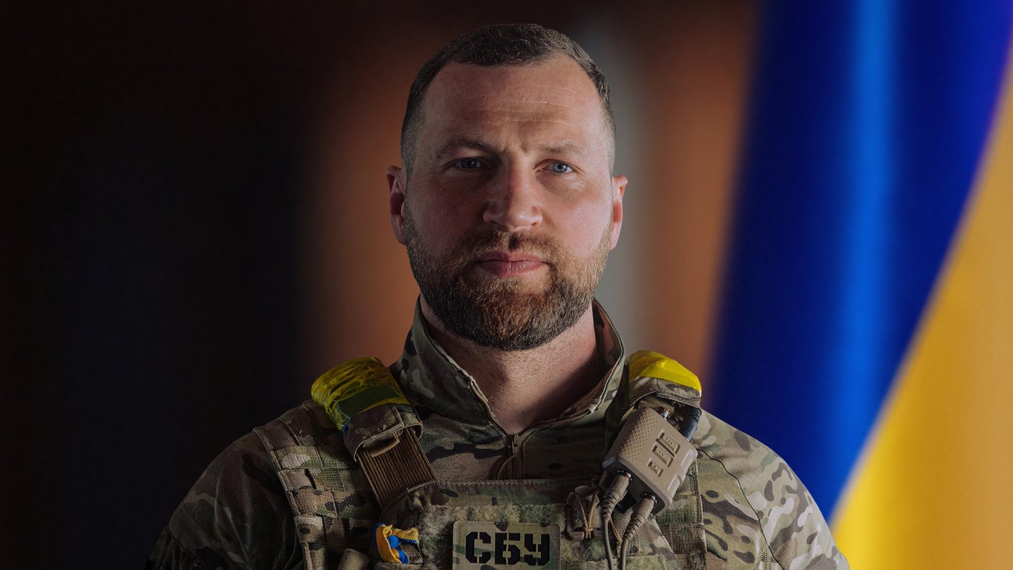 Yevhenii Khmara in military uniform standing in front of a Ukrainian flag