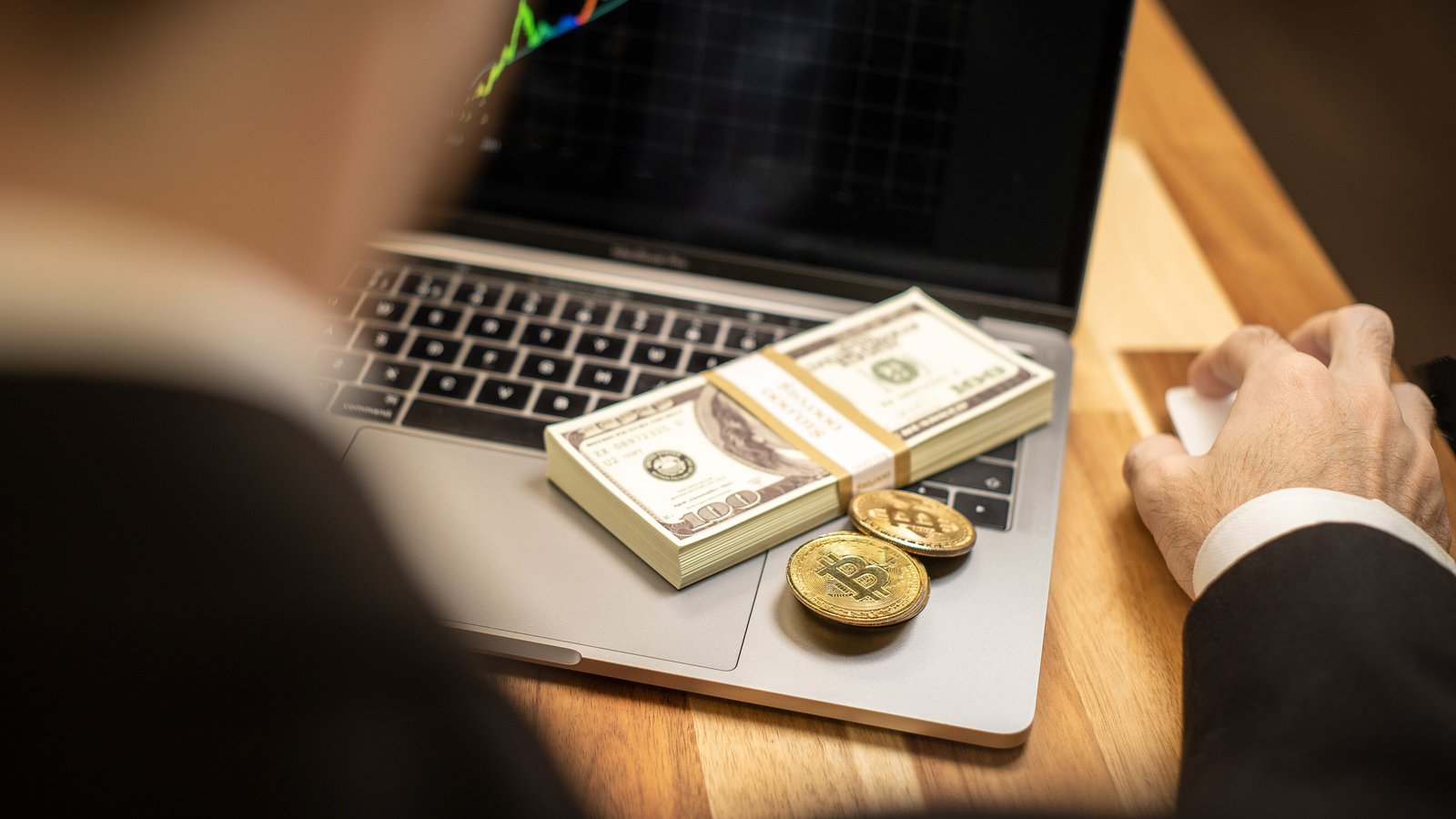 Bitcoin-style coins and a stack of US$100 notes on a laptop keyboard, with a price chart on the screen