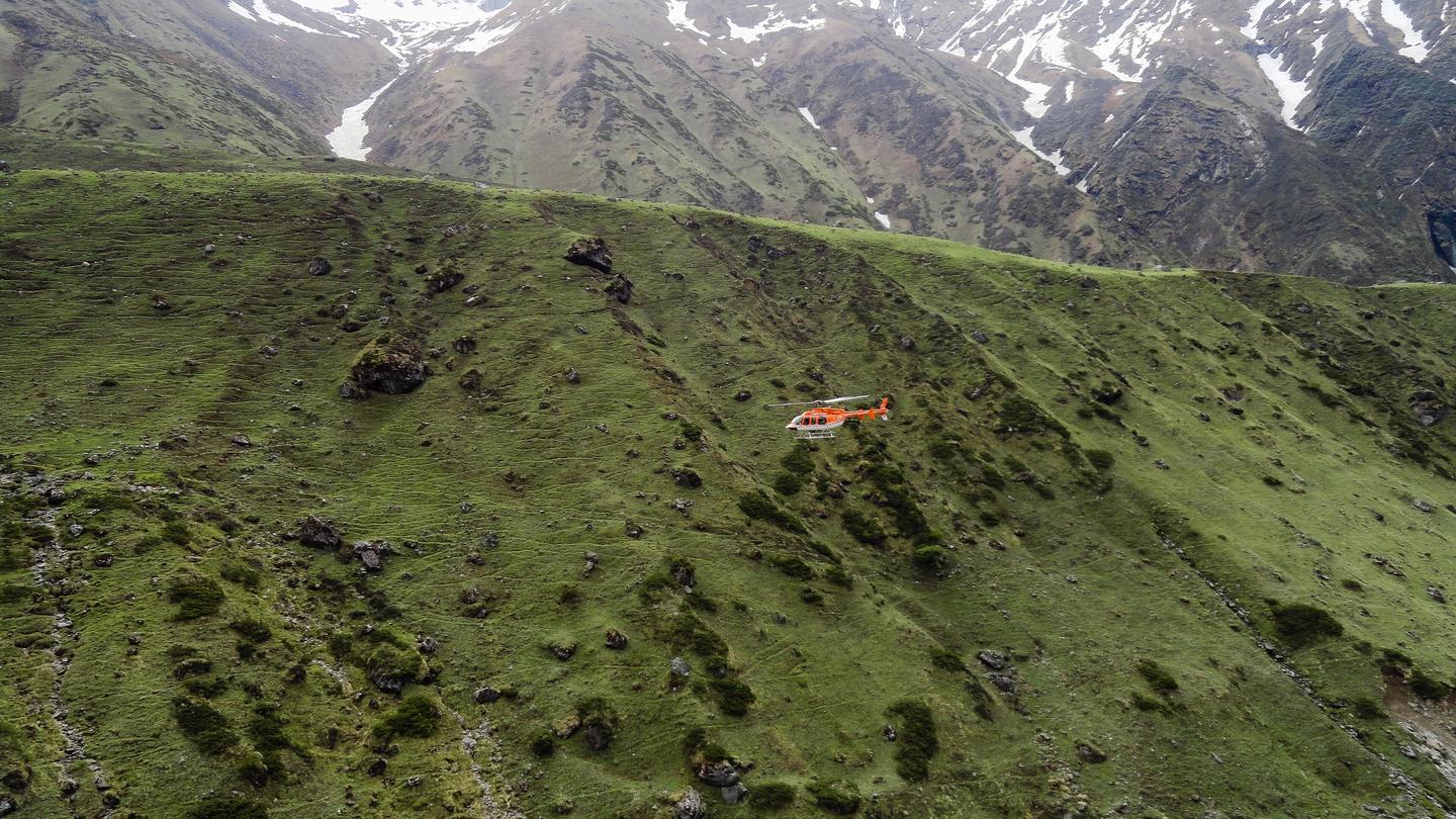 An orange helicopter flying low over a green Himalayan hillside, with snow-streaked mountains behind