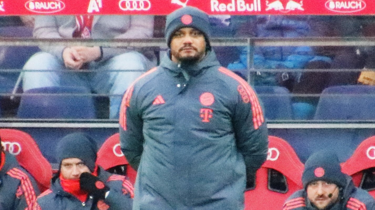 Bayern Munich head coach Vincent Kompany standing in front of the team bench in a grey Bayern coat and hat, with coaching staff seated behind him