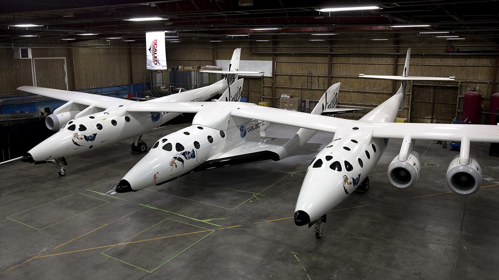 Virgin Galactic's white twin-fuselage carrier aircraft Eve and a white SpaceShipTwo spaceplane parked inside a hangar