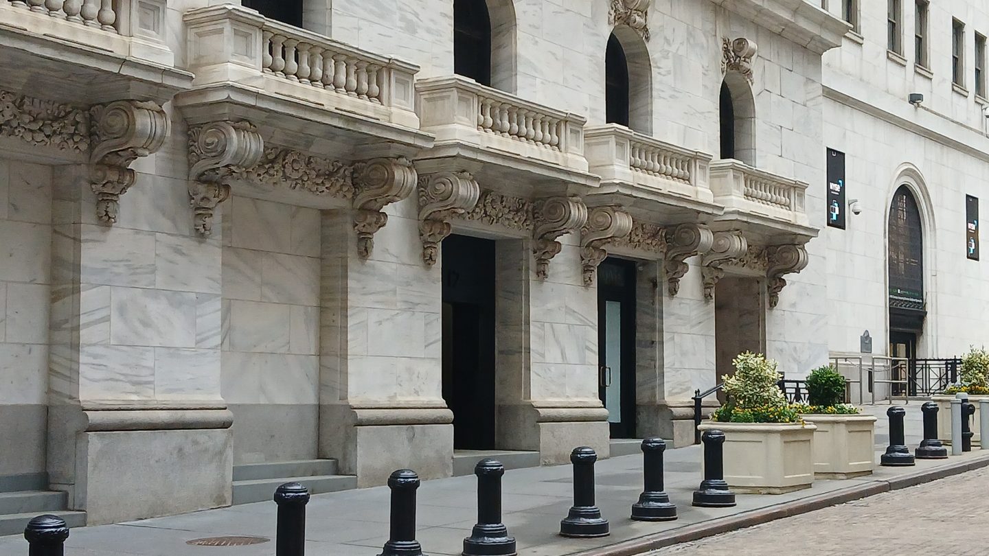 The marble Wall Street side of the New York Stock Exchange building, with NYSE banners, balconies and black bollards on the pavement