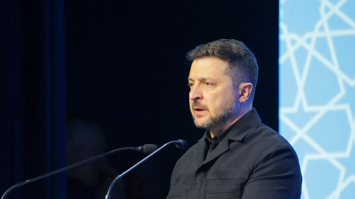 Ukrainian President Volodymyr Zelensky, in a dark jacket, speaking at a microphone in front of a blue patterned backdrop